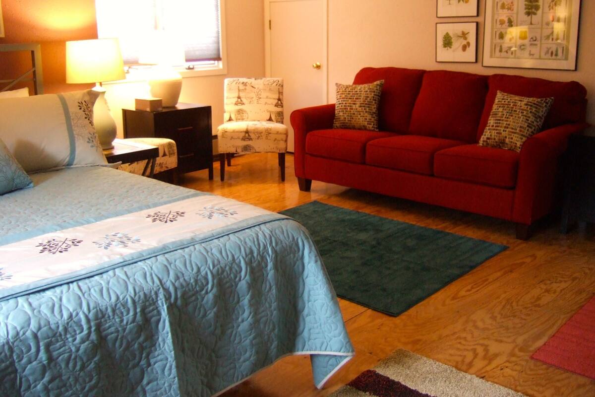 A room with a bed, a red sofa, a chair, a table, and a lamp.