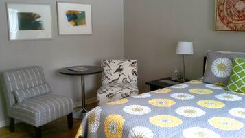 A hotel room with a grey patterned armchair, a round table, a floral bedspread, a nightstand with a lamp, and two framed artworks on the wall.