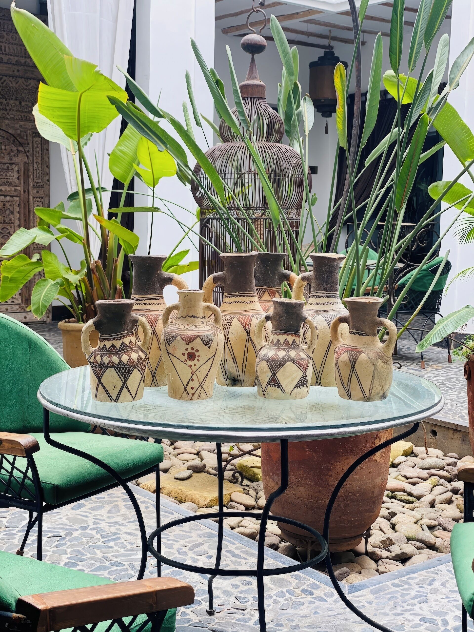 A round glass table with decorative pottery, surrounded by green chairs and potted plants.