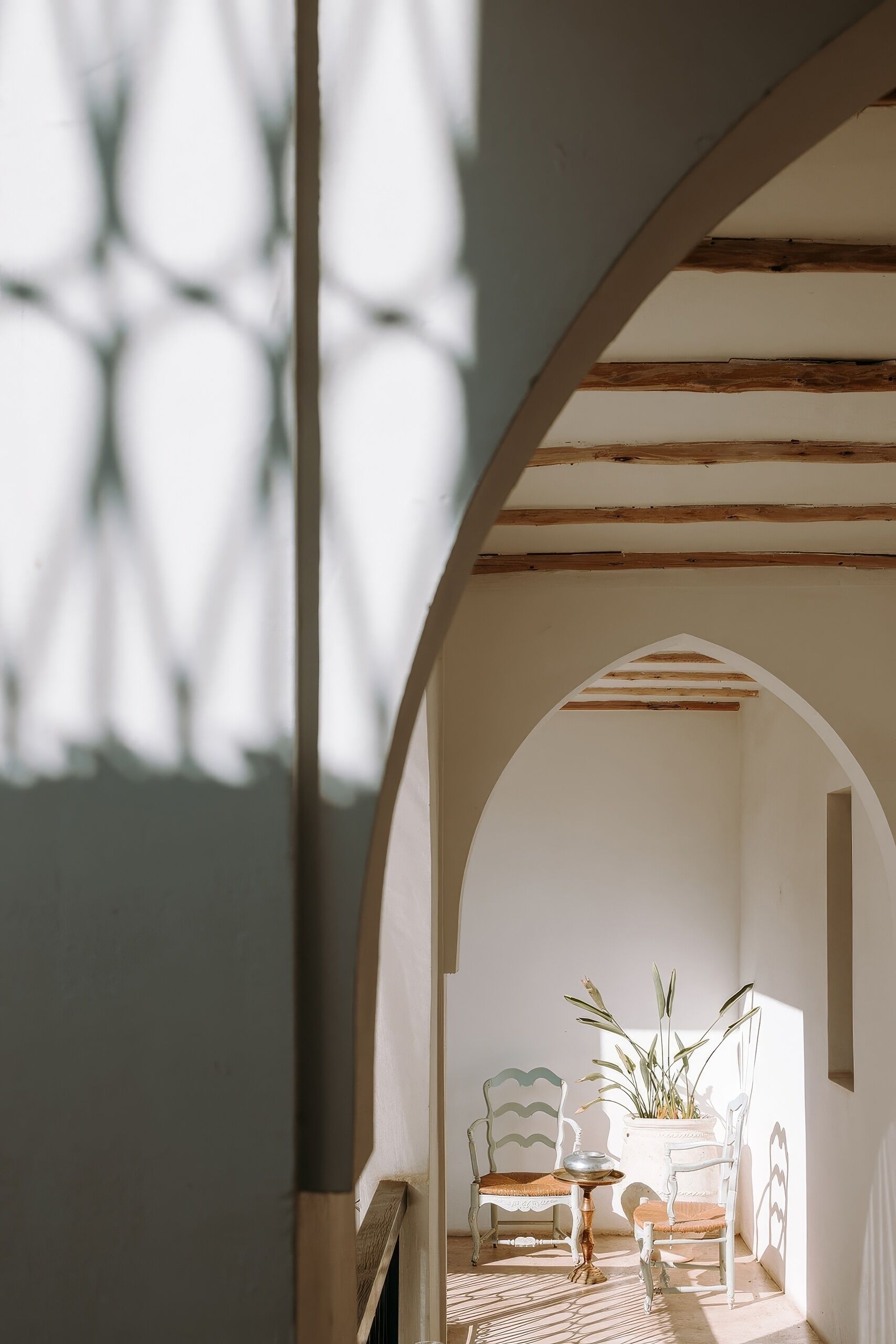 Un arco de entrada a una habitación iluminada por el sol, con dos sillas, una mesa, una planta en maceta y un suelo con diseño.
