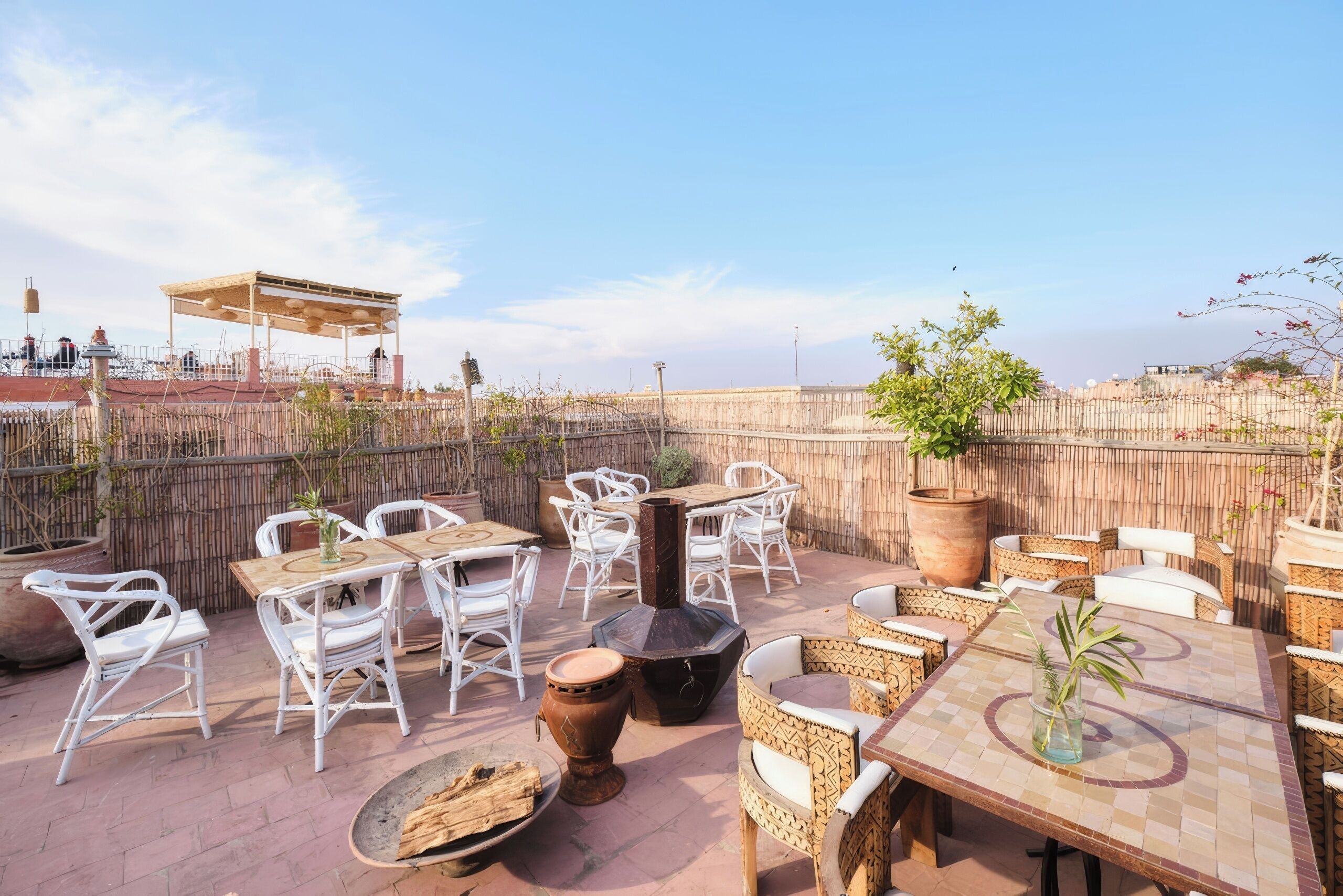 An outdoor patio with white wicker chairs and tables, a large planter, and a brick wall in the background.