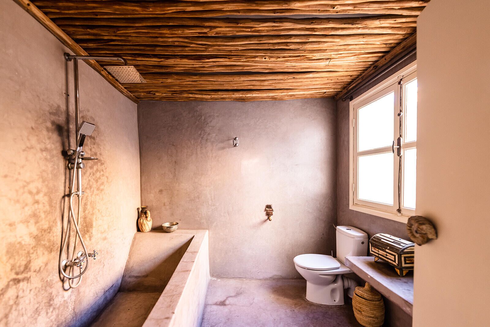 A rustic bathroom with a wooden ceiling, a bathtub, a toilet, and a window.