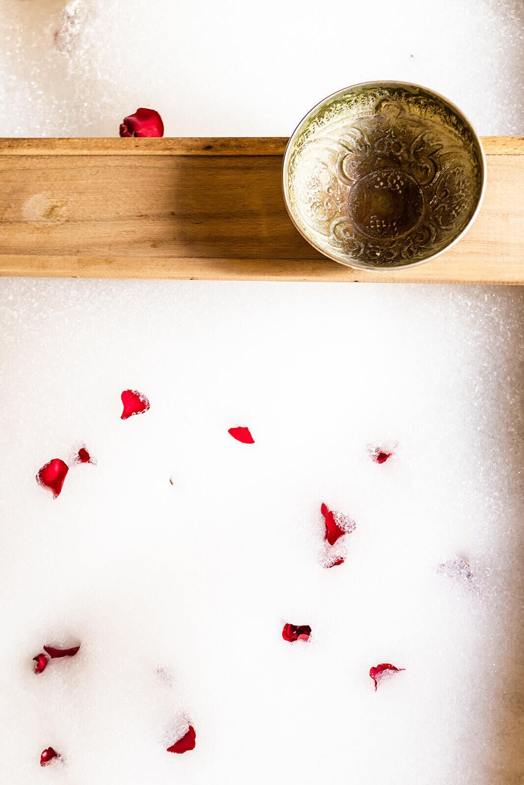 A wooden tray with a decorative bowl on it, placed on a white surface with scattered red rose petals.