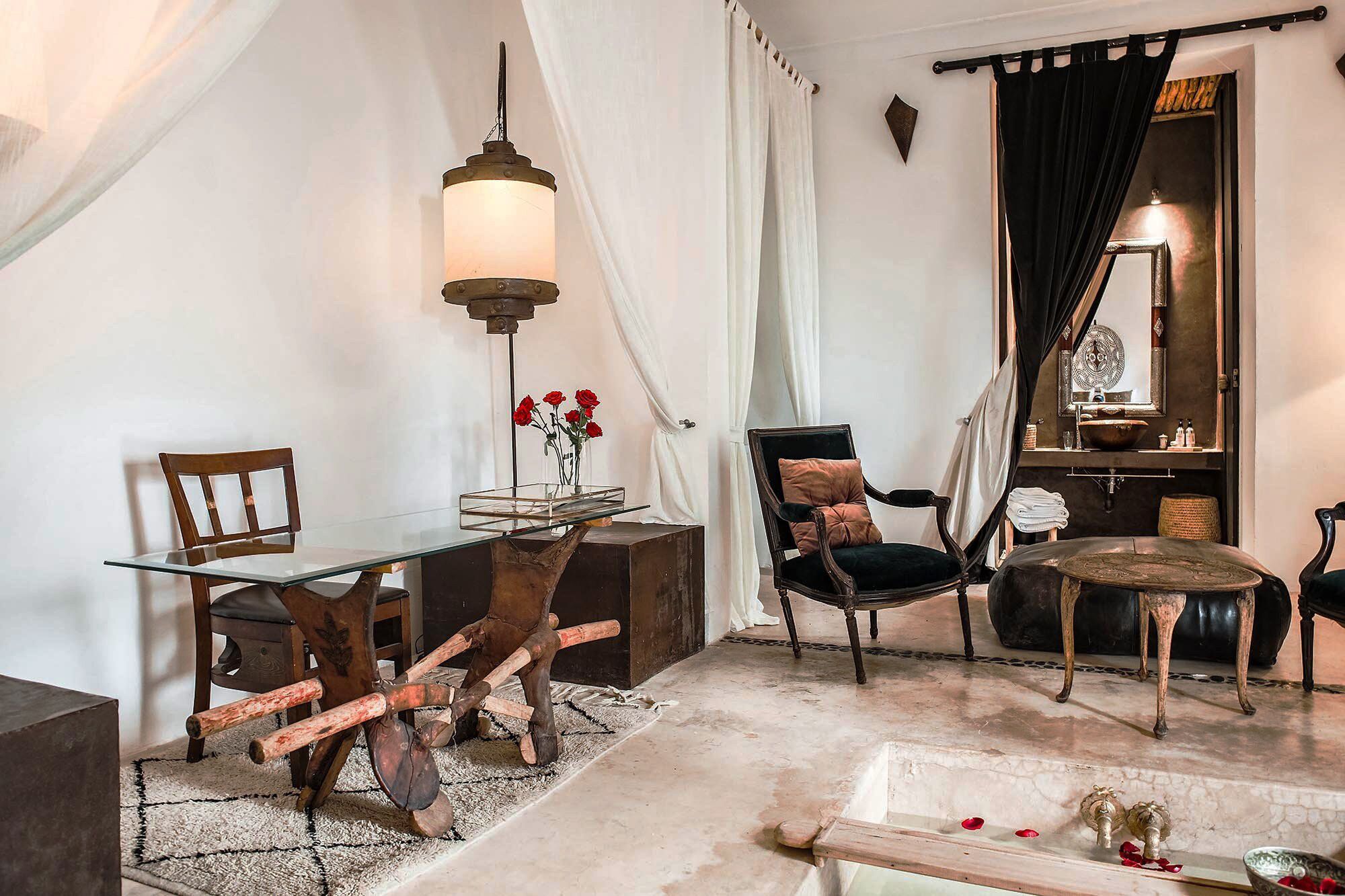 Una habitación acogedora con una mesa de vidrio, una silla de madera, un sillón tapizado negro, una mesa redonda y una lámpara colgante.