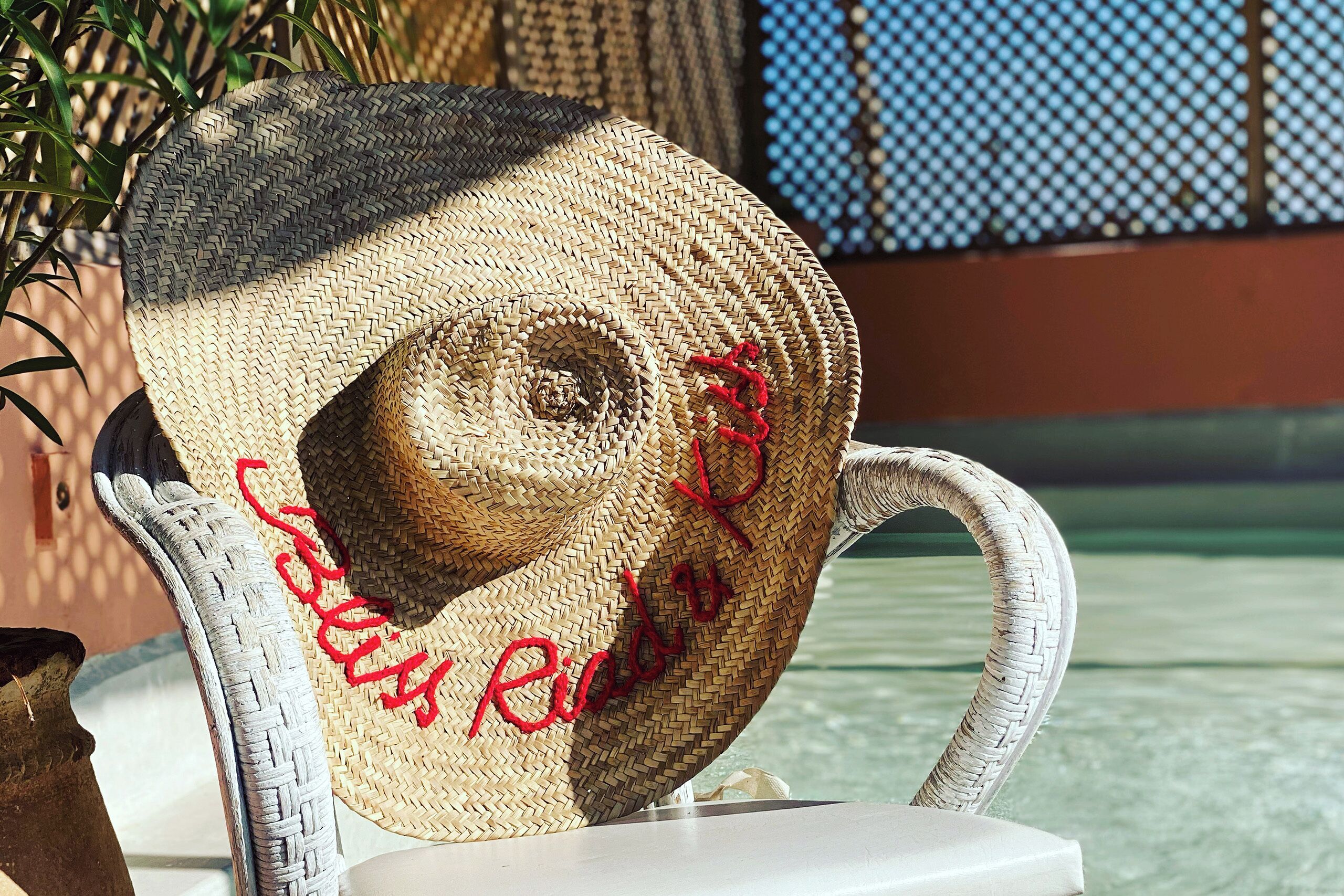 A straw hat with red embroidery on a white chair by a poolside.