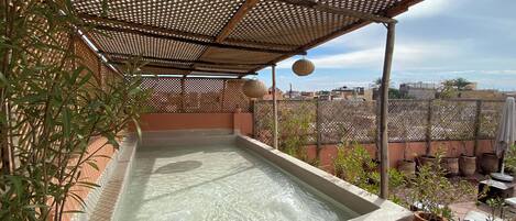 A rooftop hot tub with a wooden pergola overhead, surrounded by potted plants and a view of the cityscape.