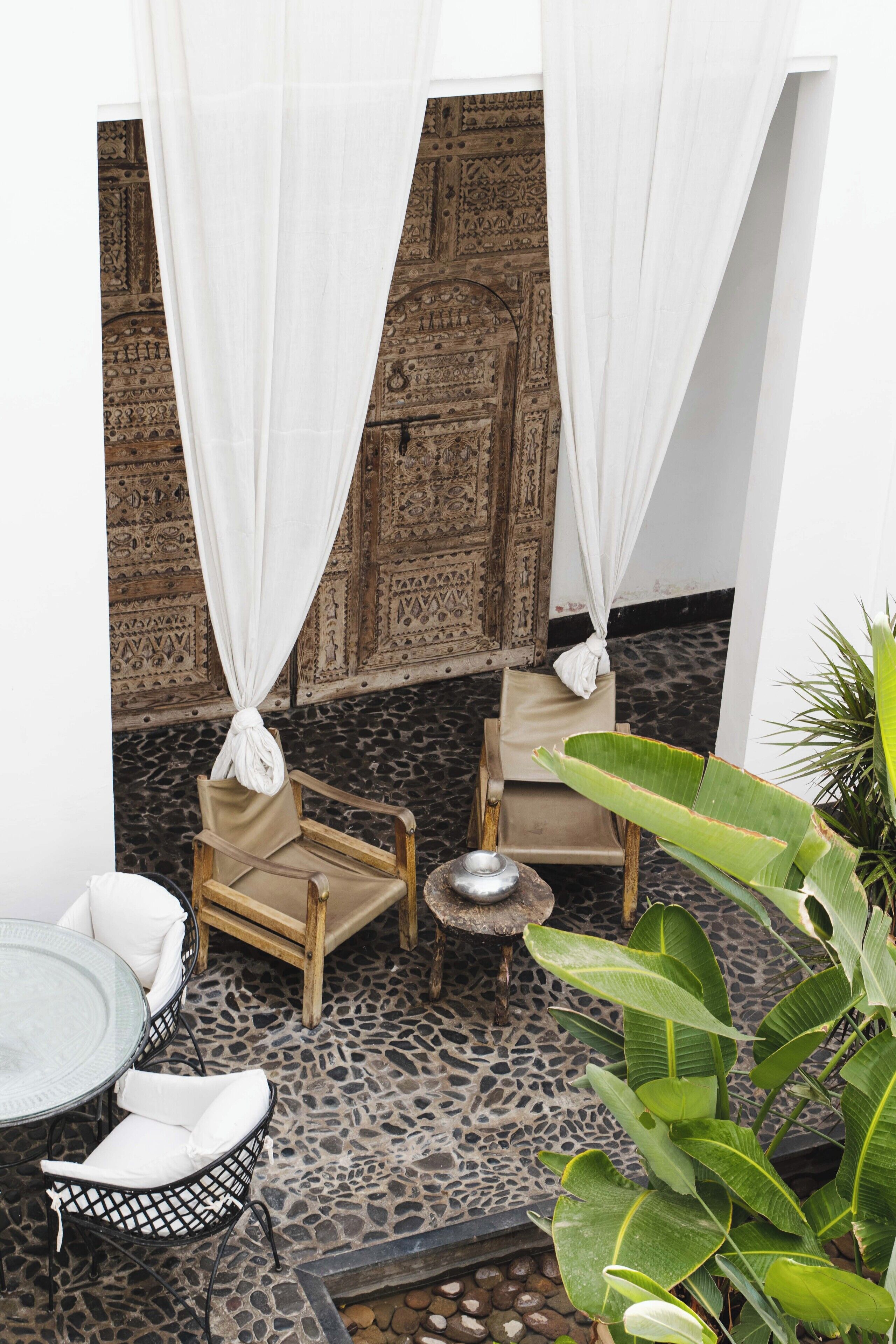 An outdoor seating area with two wooden chairs and a small table, surrounded by a patterned wall and a potted plant.