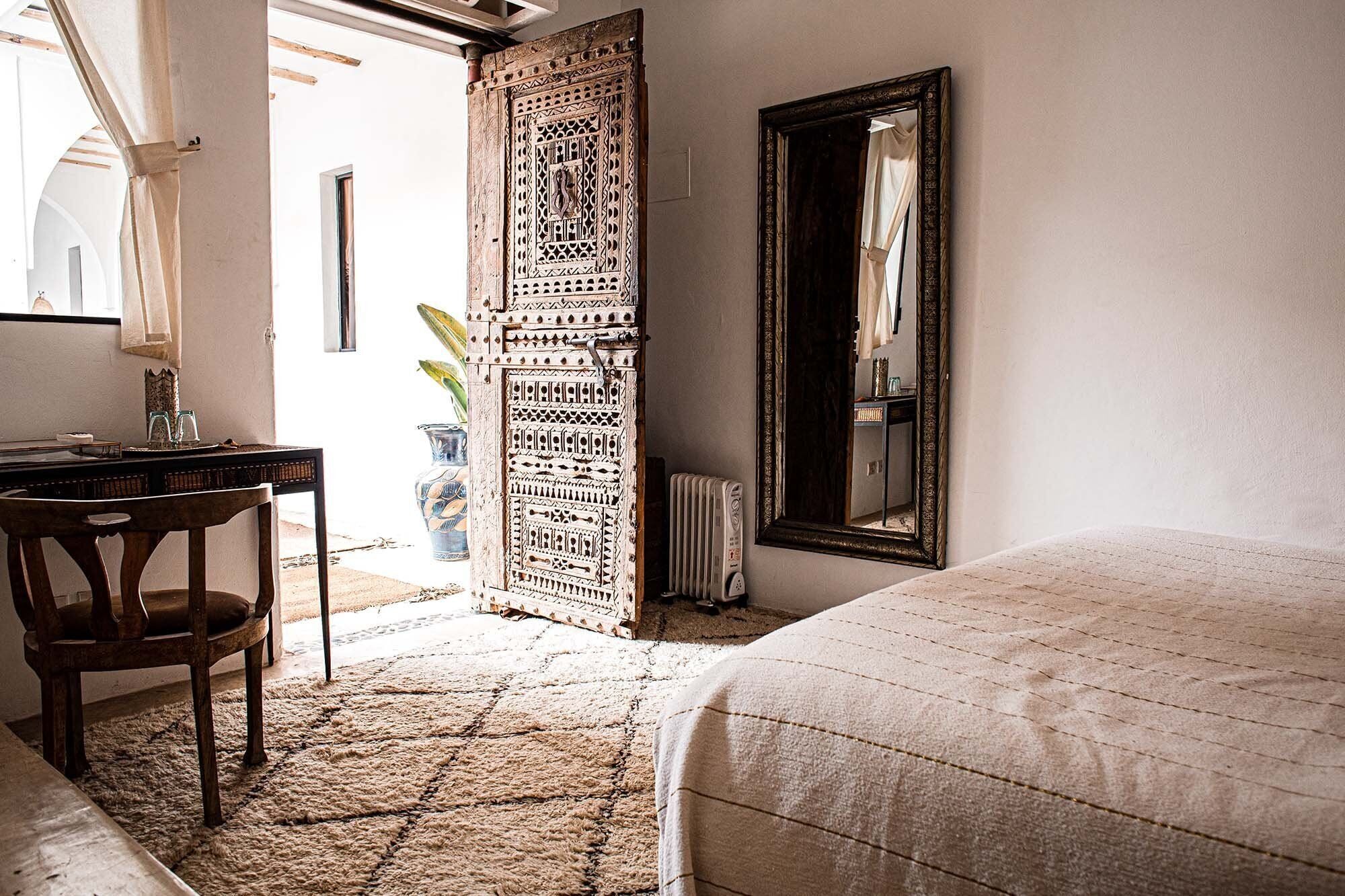 A bedroom with a bed, a wooden chair, a desk with a mirror, and a decorative door.
