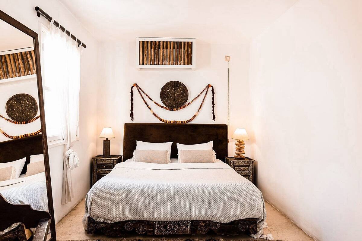 A bedroom with a large bed, two bedside tables, a mirror, and a window with curtains.