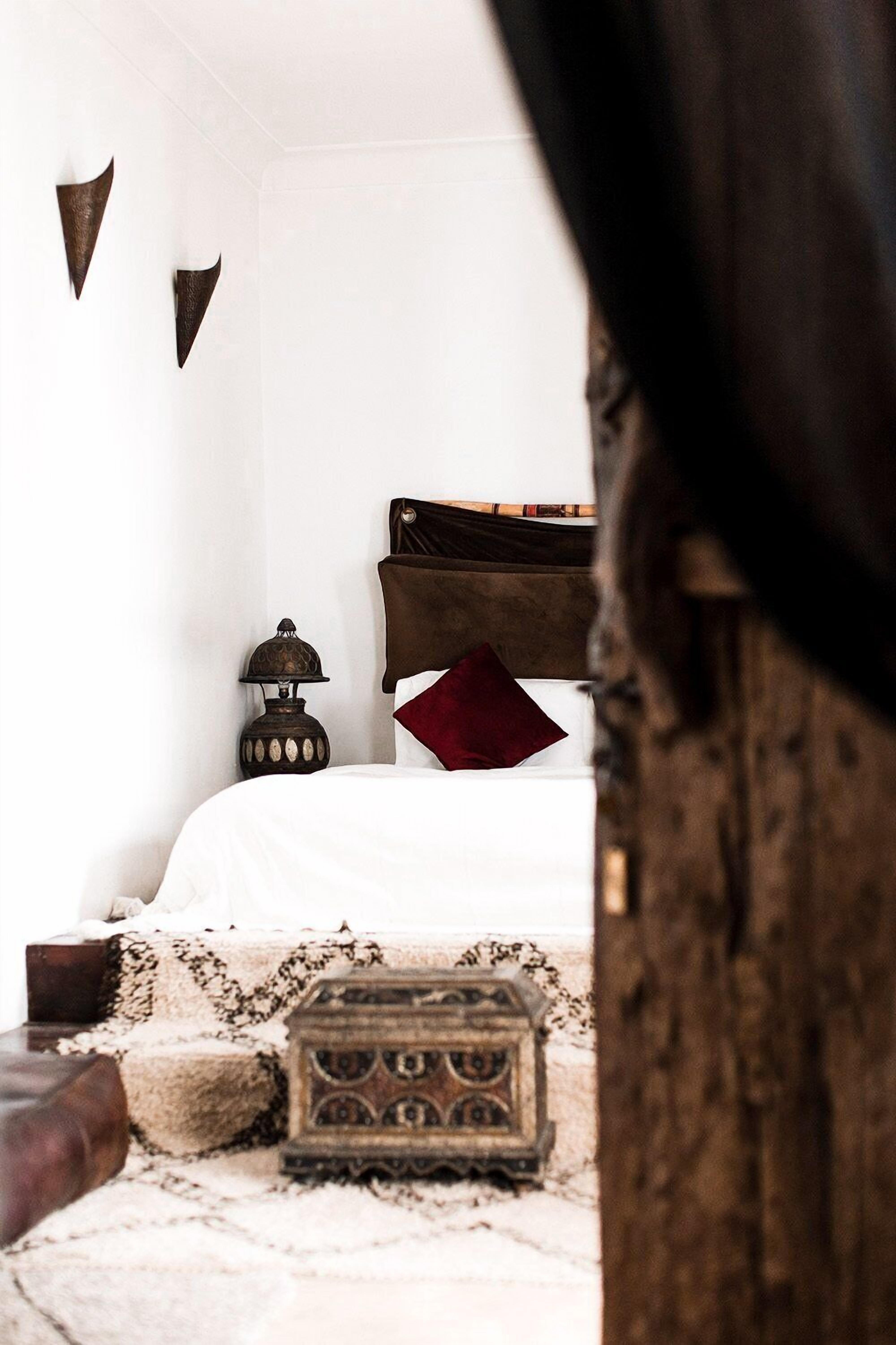 Un dormitorio con cama, un cofre de madera, una lámpara y una estantería en la pared.