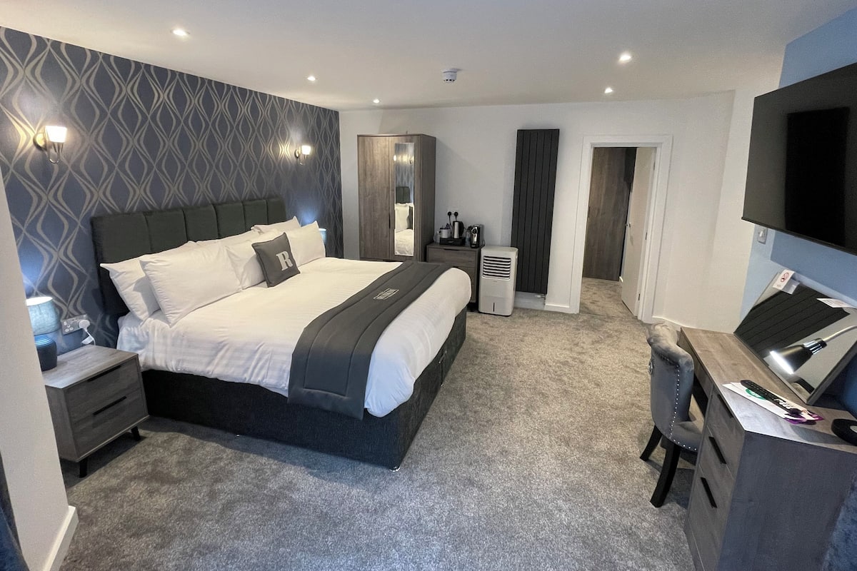 A modern hotel room with a large bed, a desk with a computer, a flat-screen TV, and a walk-in closet.