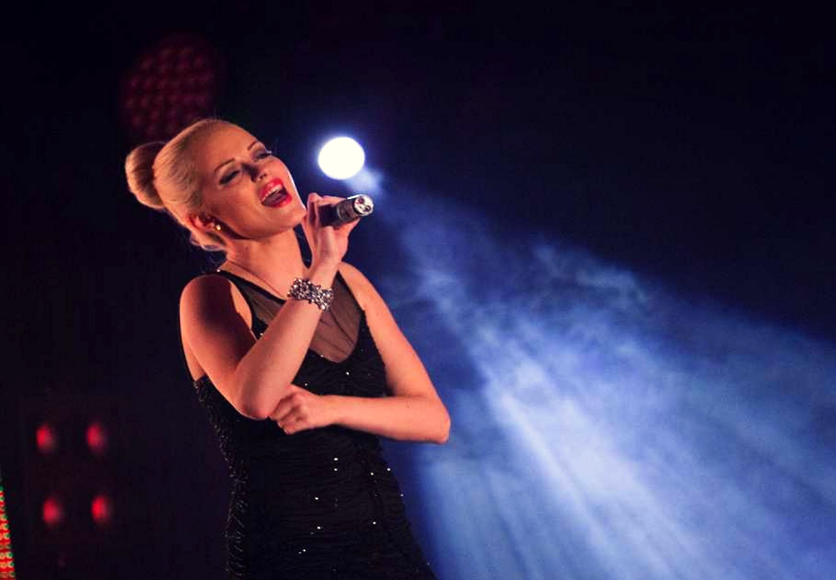 A person in a black dress holding a microphone, with stage lighting in the background.