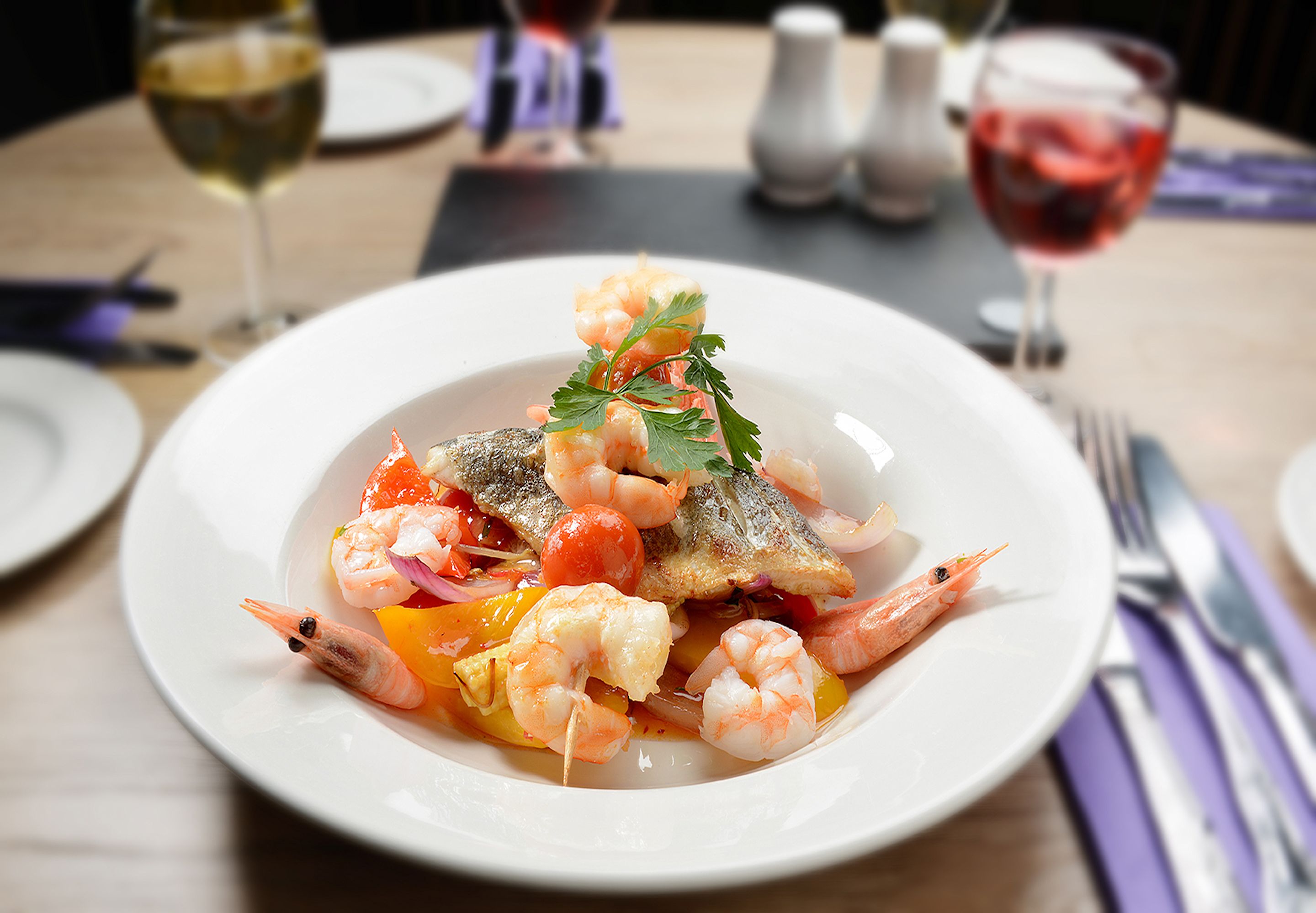 A plate of seafood dish with a fish, shrimp, and vegetables, accompanied by wine glasses and cutlery on a table.