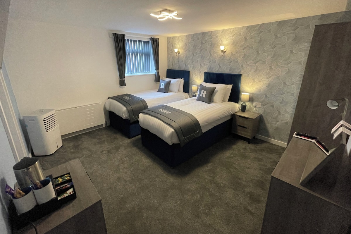 A hotel room with two beds, a large window with curtains, a wall-mounted light fixture, and a bedside table with a lamp.