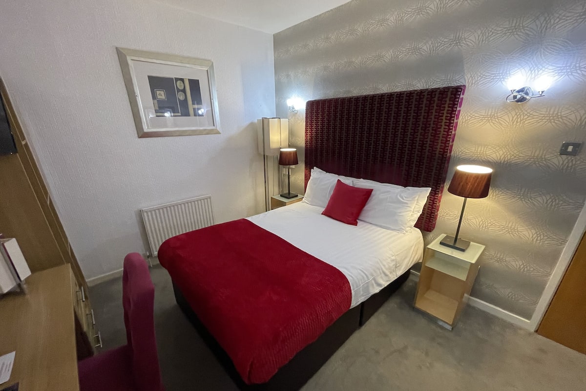A hotel room with a large bed, a red throw blanket, two bedside lamps, a framed picture on the wall, and a radiator.