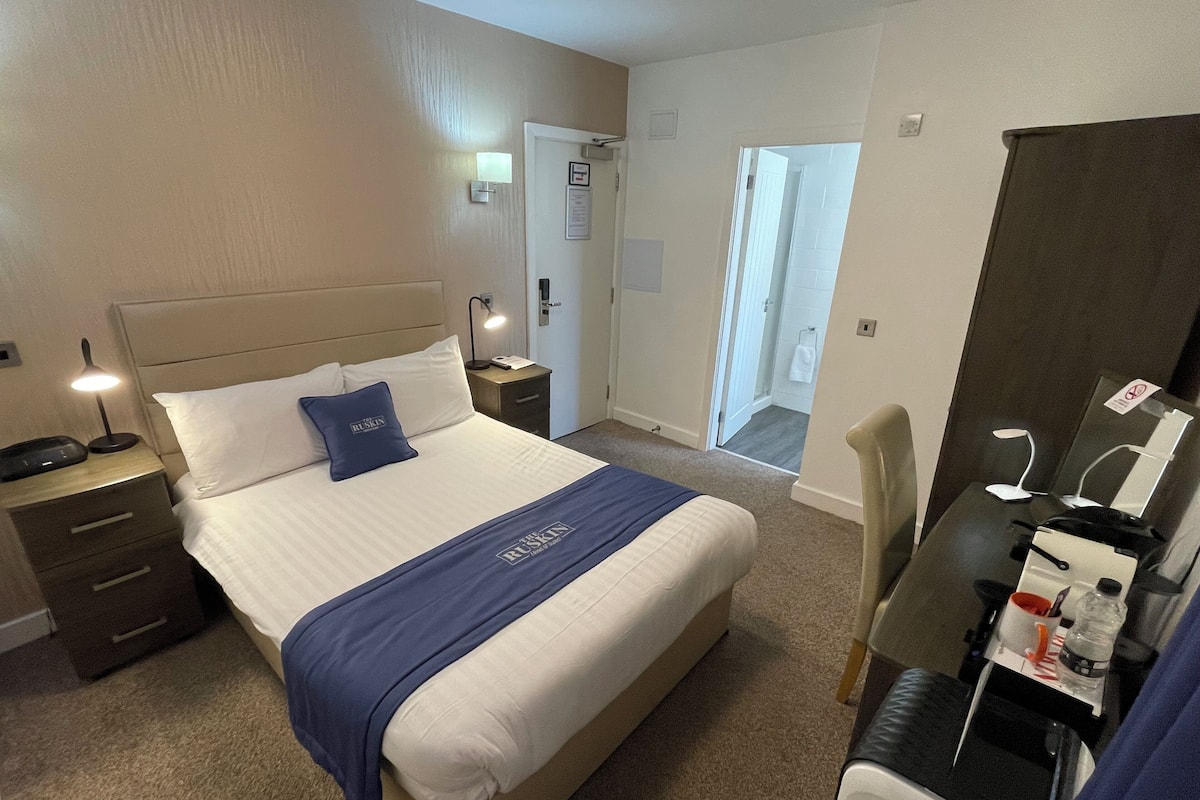 A hotel room with a bed, bedside tables, a desk with a chair, a mirror, and a bathroom visible through an open door.
