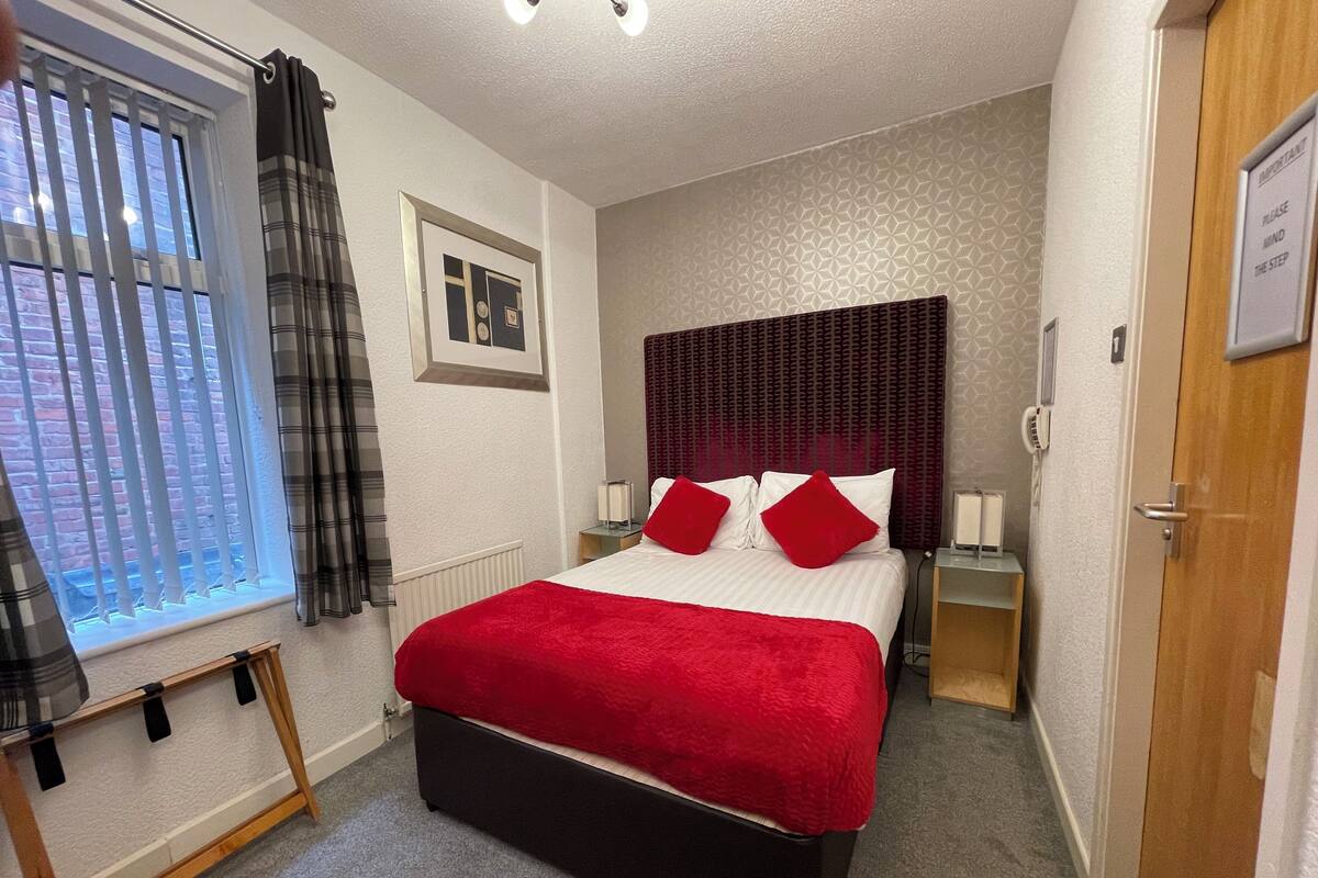 A hotel room with a bed, a window with blinds, a bedside table with a lamp, and a door with a sign.