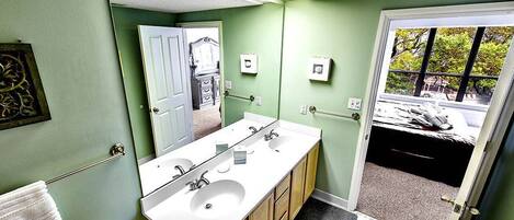 A bathroom with a double sink, a toilet, and a mirror. There is a towel rack and a door leading to a bedroom.