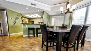 A dining area with a wooden floor, a marble table, and black chairs. There is a kitchen in the background with a green wall and a decorative plant. - Sunset Escape 2 Bedroom Holiday Home (Hilton Head Island)