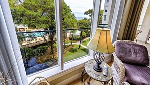 A room with a large window overlooking a pool and greenery. There is a wicker chair and a small table with a lamp. - Sunset Escape 2 Bedroom Holiday Home (Hilton Head Island)