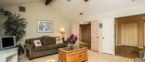 A spacious living room with a ceiling fan, a television, a sofa, a coffee table with flowers, and a large plant.