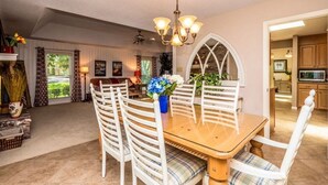 Un comedor con mesa de madera y sillas blancas, una lámpara de araña y vista a la cocina.