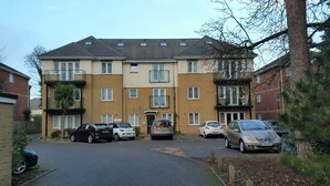 Um edifício residencial de vários andares com entrada central, várias janelas e um estacionamento com diversos carros.