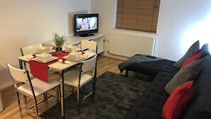 Uma sala de estar moderna com área de jantar, um sofá preto com almofadas vermelhas, uma televisão e uma janela com persianas.