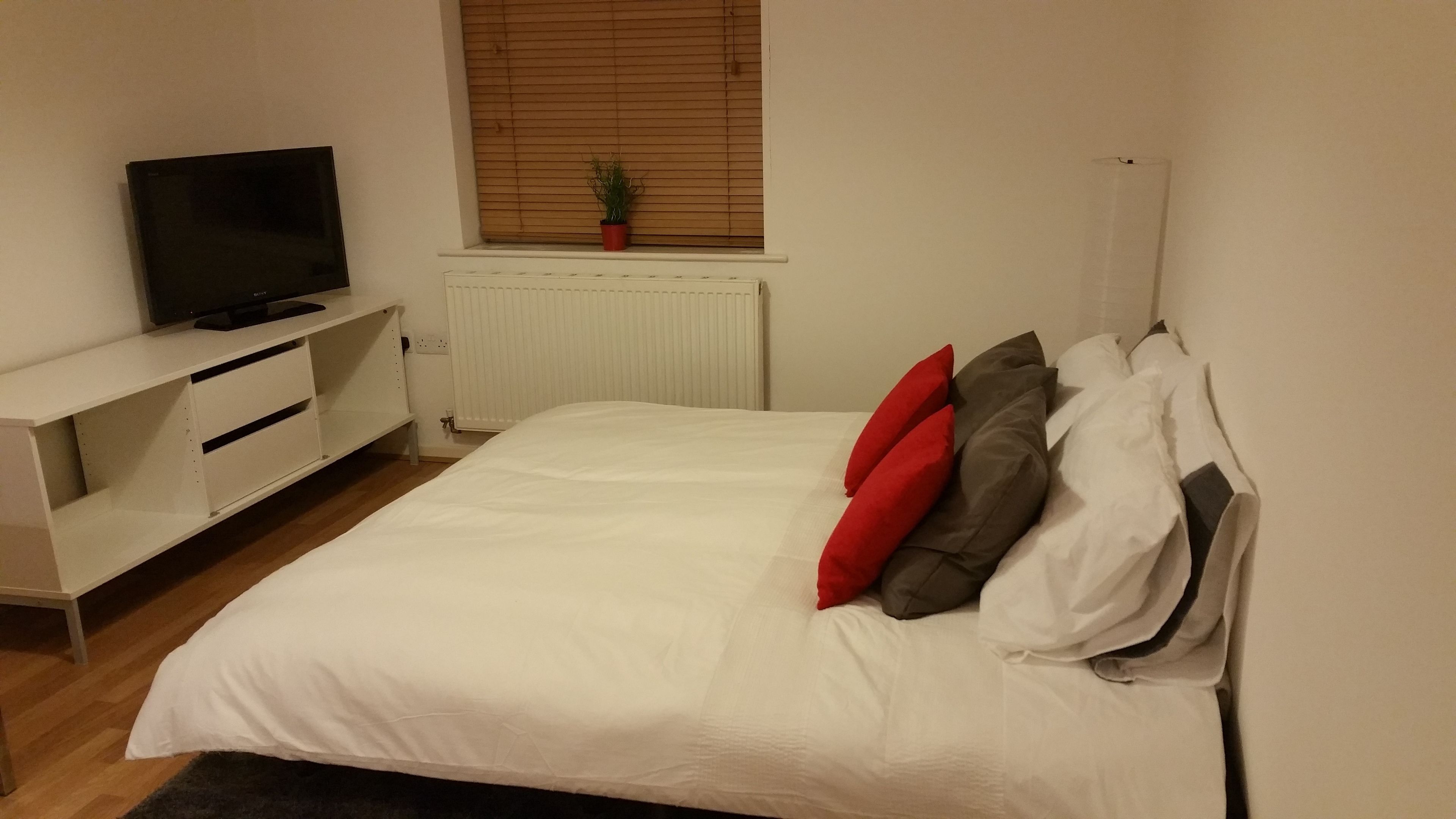 A bedroom with a bed, a TV on a white stand, a window with blinds, and a radiator.