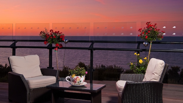 Une terrasse dotée de meubles en osier et d’une table, offrant une vue sur le coucher de soleil sur la mer.