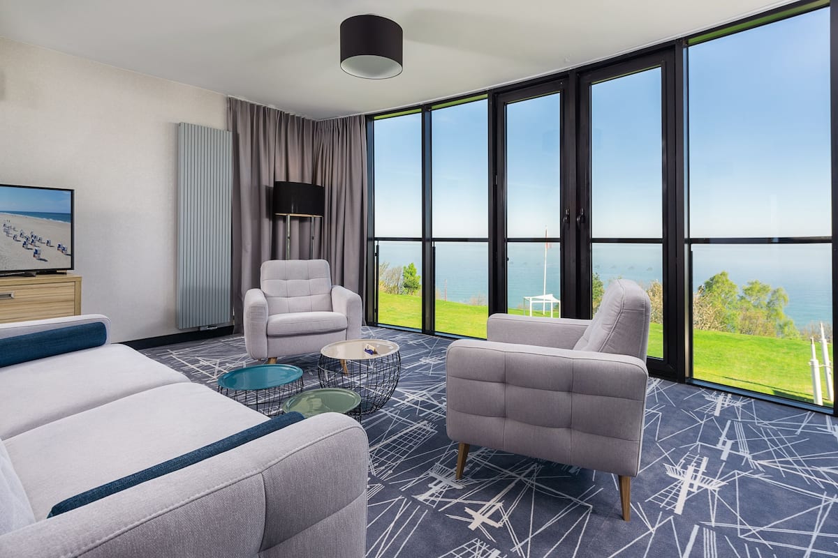 Une chambre d’hôtel moderne dotée d’une grande fenêtre donnant sur la mer, d’un canapé gris, d’un canapé blanc, d’une table basse ronde et d’une lampe.