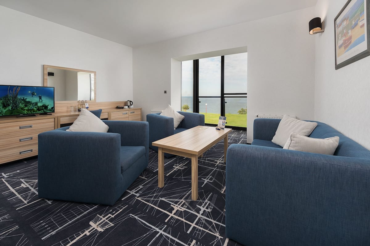 Une chambre d’hôtel moderne dotée d’une grande fenêtre donnant sur la mer, d’un téléviseur à écran plat, d’une table basse en bois et de deux canapés bleus.