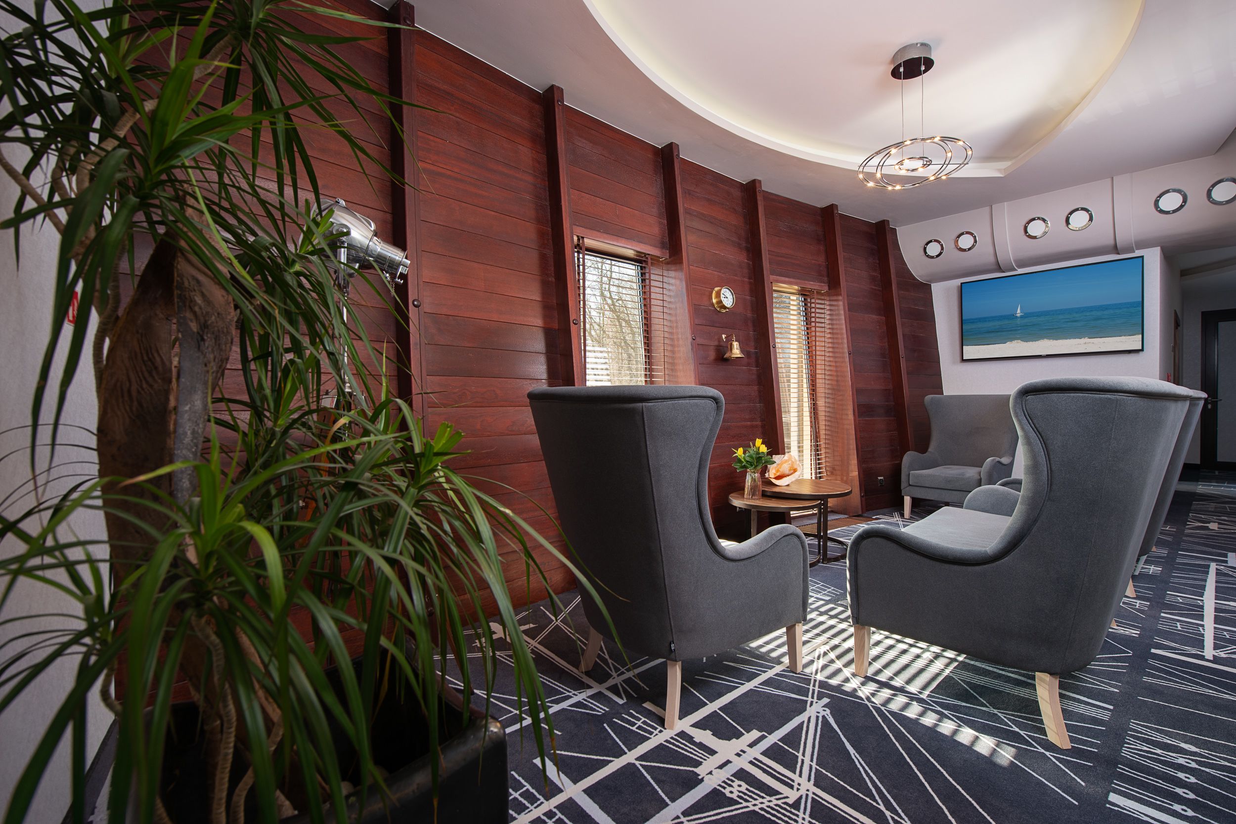 Una habitación de hotel moderna con paneles de madera oscura, dos sillones tapizados de gris, una mesa redonda con un jarrón y un cuadro grande que representa una playa.