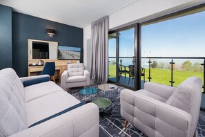 Une chambre d’hôtel moderne dotée d’un balcon, d’un canapé, d’un fauteuil, d’un bureau et d’une grande fenêtre donnant sur l’extérieur.