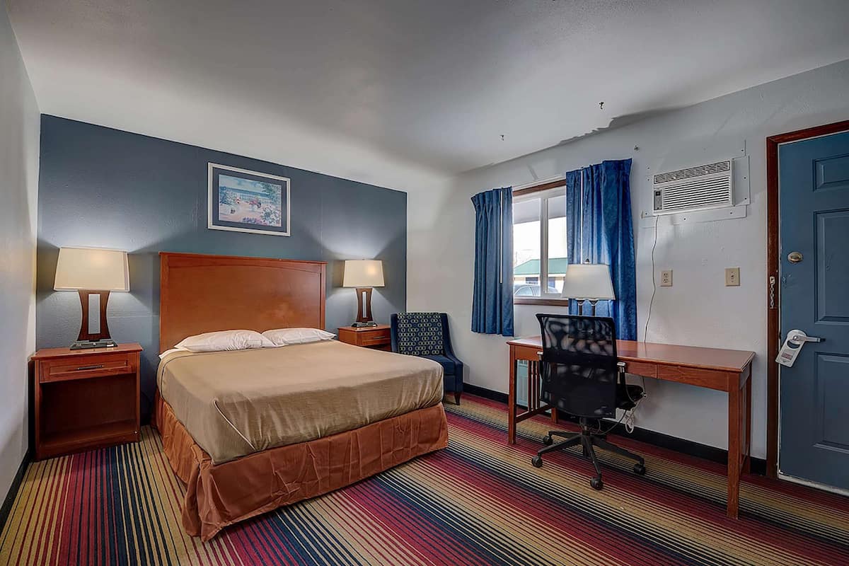A hotel room with a bed, two bedside tables with lamps, a desk with a chair, a window with curtains, and a door.