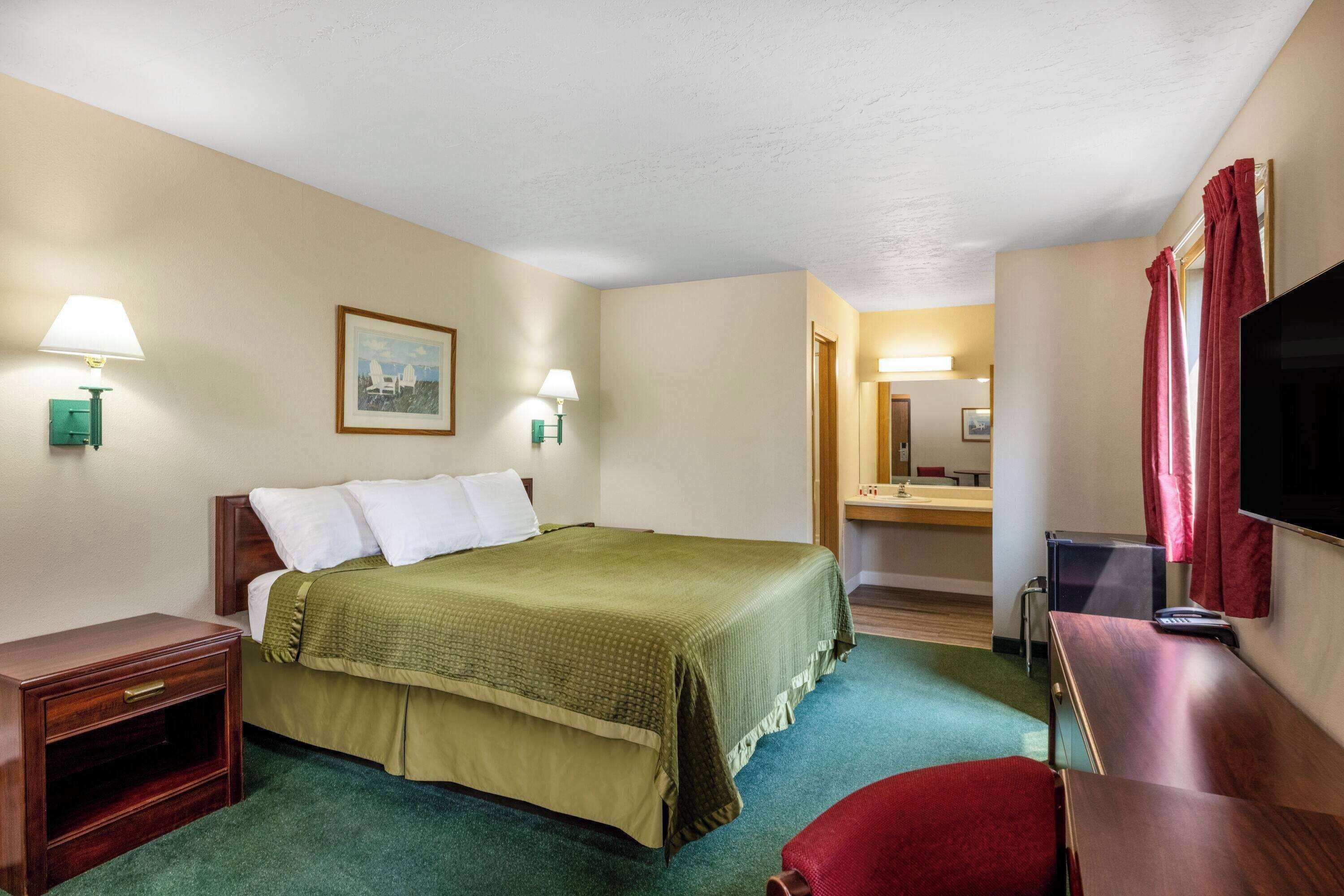 A hotel room with a bed, bedside tables, a desk, a television, and a bathroom visible through an open door.
