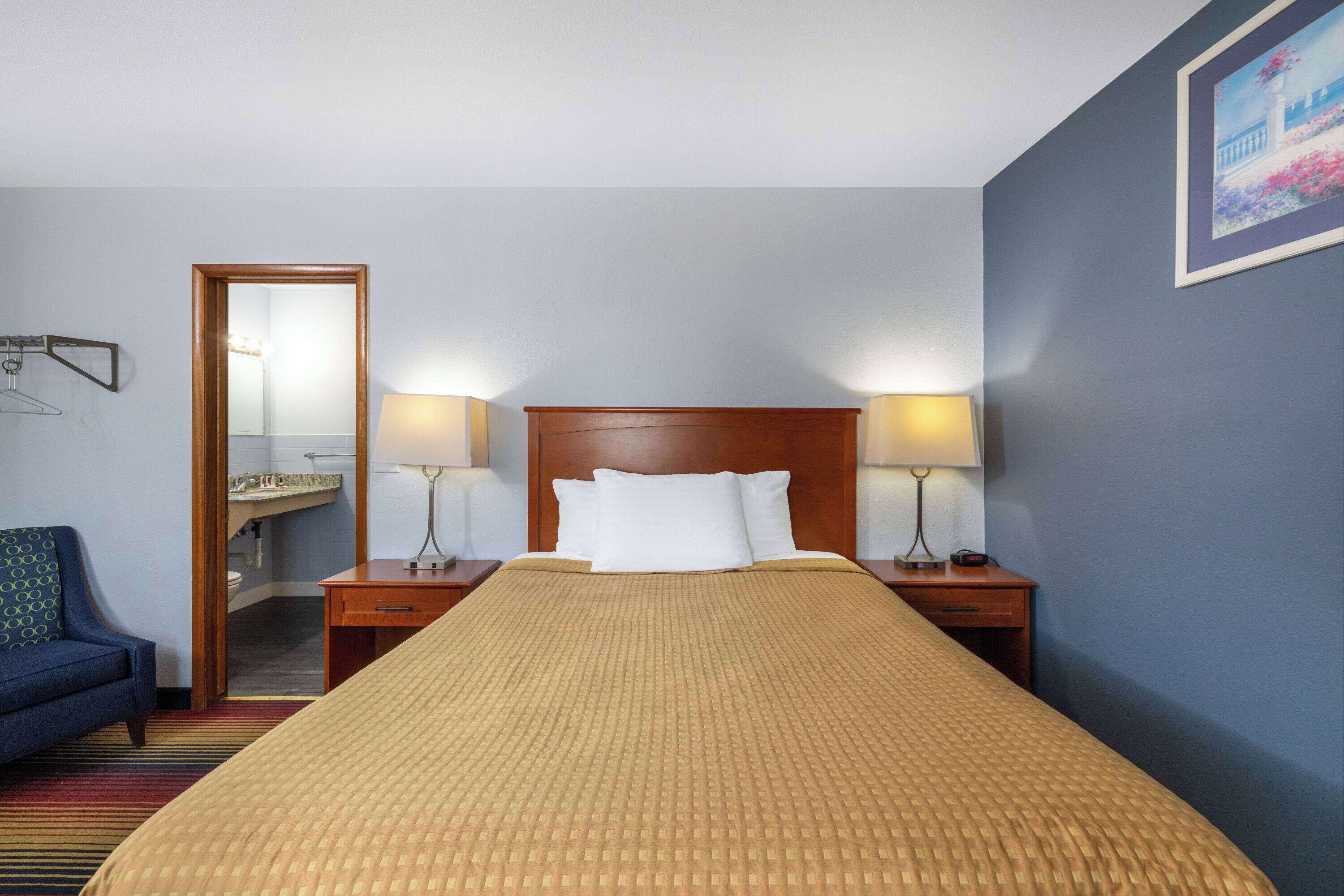 A hotel room with a large bed, two bedside tables with lamps, a wooden headboard, a blue armchair, and a mirror on the left wall.