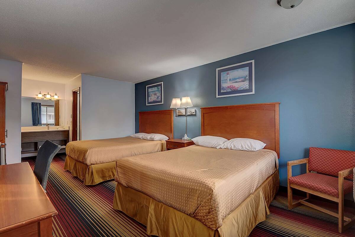 A hotel room with two beds, a desk, a chair, and a lamp.