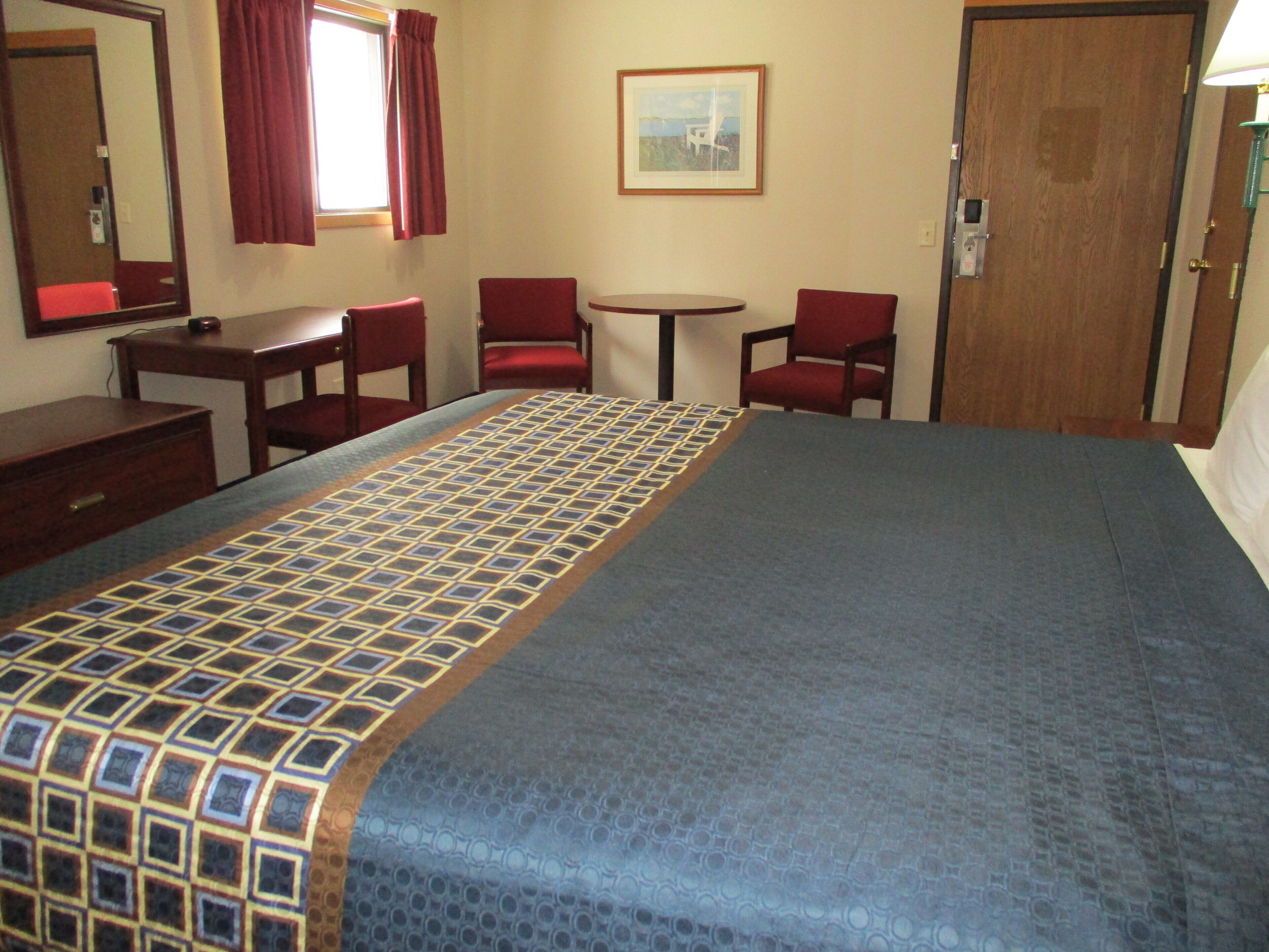 A hotel room with a bed, two red chairs, a small table, a mirror, a window with red curtains, and a door.
