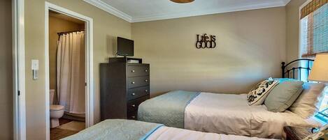 Een slaapkamer met twee bedden, een plafondventilator, een dressoir, een televisie en een badkamer die door een open deur zichtbaar is.