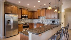 A modern kitchen with wooden cabinets, stainless steel appliances, a granite countertop, and bar stools. - Acqualina 9 Bedroom Holiday Home by Five Star Properties (Miramar Beach)