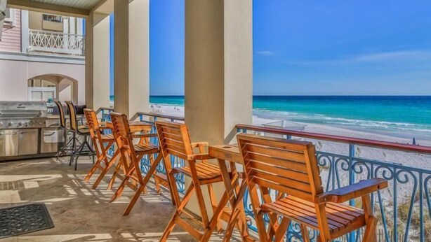 Un espace extérieur aménagé, abrité et doté de chaises en bois et d’un barbecue, offrant une vue sur une plage aux eaux d’un bleu cristallin.