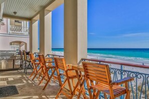 A covered outdoor seating area with wooden chairs and a grill, overlooking a beach with clear blue water. - Bella Beach 5 Bedroom Holiday Home by Five Star Properties (Miramar Beach)