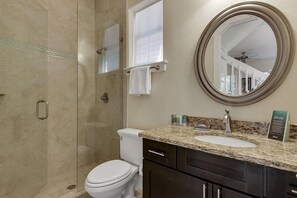 A bathroom with a glass shower enclosure, a white toilet, a sink with a granite countertop, and a round mirror. - Beyond the Sea 5 Bedroom Holiday Home by Five Star Properties (Miramar Beach)