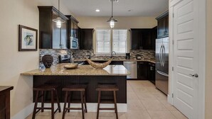 Una cocina moderna con armarios oscuros, encimera de granito, una isla grande con taburetes, microondas integrado y un refrigerador de acero inoxidable.