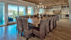 A spacious dining area with a wooden table and woven chairs, large windows offering a view of the outdoors, and a kitchen in the background.