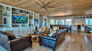 A spacious living room with a large TV, leather sofas, wooden flooring, and a ceiling fan.