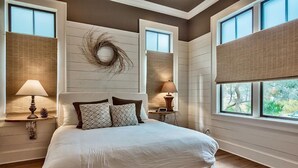 Un dormitorio con una cama grande, dos mesitas de noche con lámparas, un adorno en la pared y vistas a través de una ventana con persianas.