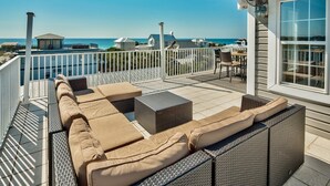 A covered outdoor seating area with a view of the ocean, furnished with a sofa and chairs, and a table.
