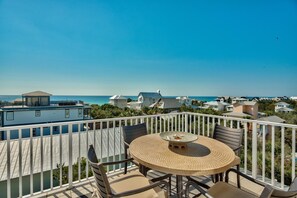 A balcony with a round table and chairs, overlooking a coastal area with houses and the sea.